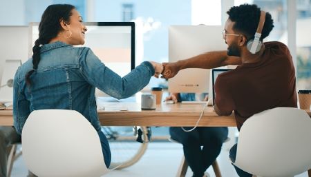 A man and woman fist bumping while working next to each other at an office desk 